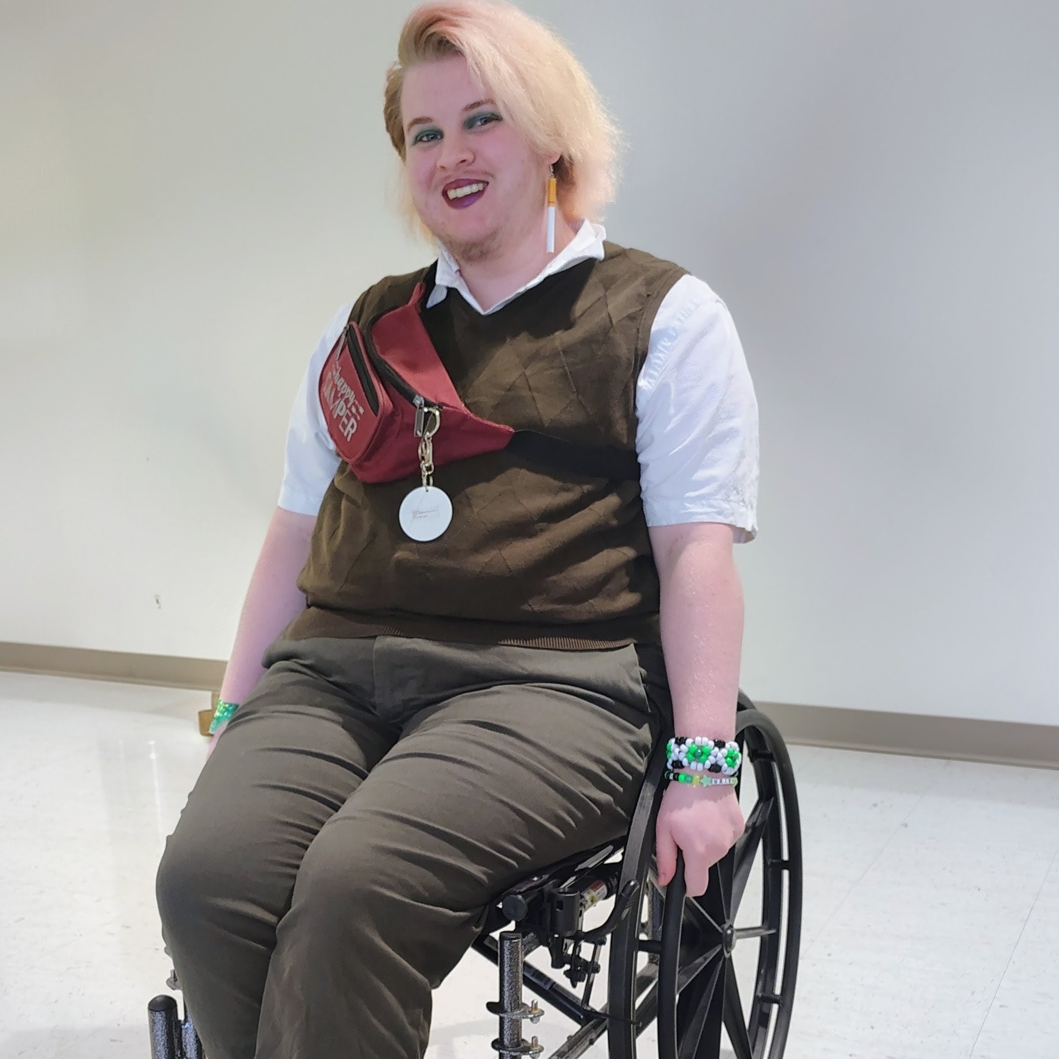 A trans man with medium length bleach blond hair is sitting in a wheelchair while smiling at the camera.